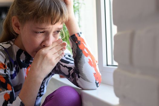 Girl weeping bitterly at the window wipes her tears with her hand
