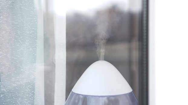 The white ultrasonic humidifier on a window of living room moistens dry air. Close-up shot