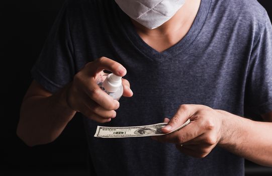 Closeup man wearing protective face mask applying sanitizer spray to 100 dollar money for cleaning, hygiene prevention COVID-19 virus or coronavirus protection concept, dark on black background