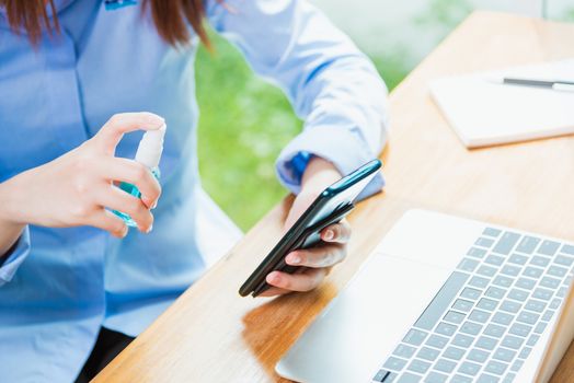 Asian young business woman wearing face mask protective working from home office with laptop computer he quarantines disease coronavirus or COVID-19 using sprays sanitizer cleaning smart mobile phone