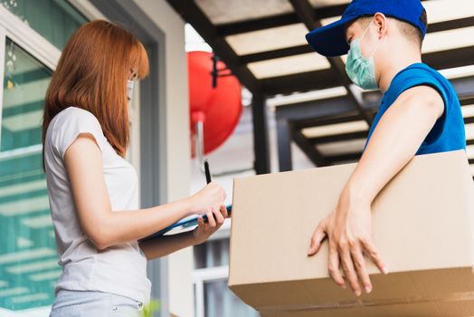 Asian young delivery man courier shopping online with package post box in uniform protective face mask coronavirus he making service woman customer using pen signature on paper book at home, COVID-19