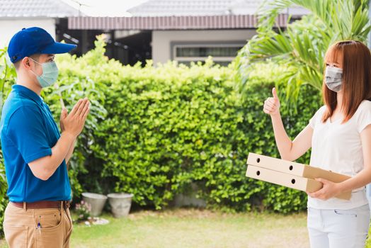 Asian delivery courier young man giving food pizza box to woman customer receive both protective face mask and show thumb up finger for good job, under curfew quarantine pandemic coronavirus COVID-19