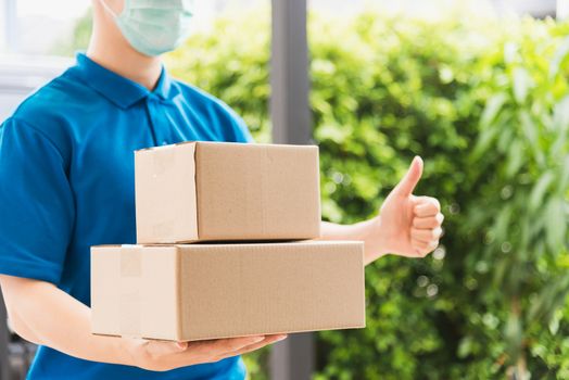 Asian delivery express courier young man use giving boxes to woman customer he wearing protective face mask at front home and thumb up finger, under curfew quarantine pandemic coronavirus COVID-19