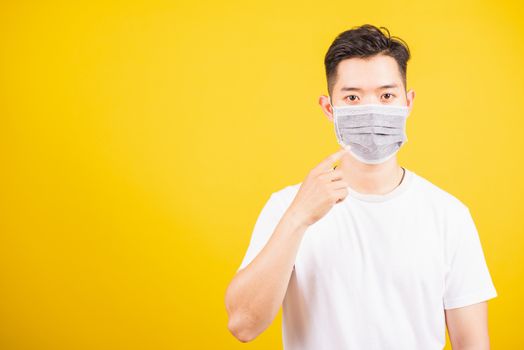 Asian young teen man wearing surgical hygienic protective face mask against coronavirus and pointing finger to mask on face, studio shot isolated white background, COVID-19 medical concept