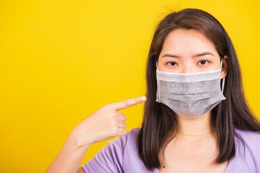 Asian young teen woman wearing surgical hygienic protective face mask against coronavirus and pointing finger to mask on face, studio shot isolated white background, COVID-19 medical concept