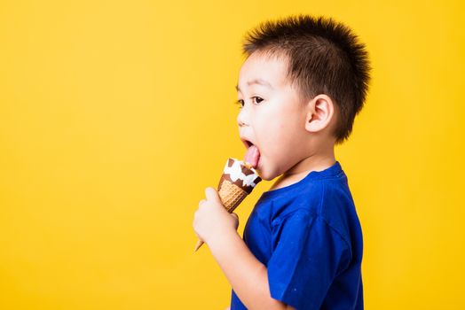 Happy portrait Asian child or kid cute little boy attractive laugh smile playing holds and eating sweet chocolate ice cream waffle cone, studio shot isolated on yellow background, summer concept