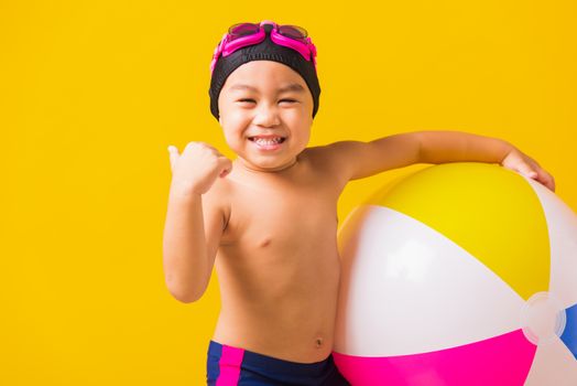 Summer vacation concept, Portrait Asian happy cute little child boy smiling in swimsuit hold beach ball and showing finger thumb up for good sign, studio shot isolated yellow background