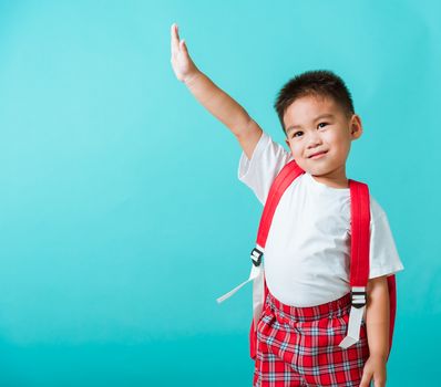 Back to school. Portrait happy Asian cute little child boy in uniform smile raise hands up glad when go back to school, isolated blue background. Kid from preschool kindergarten with school backpack