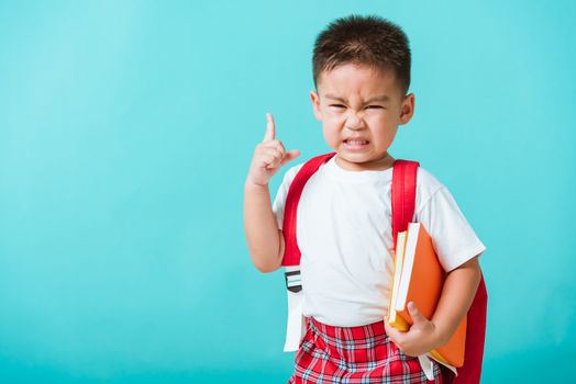 Back to school concept. Portrait Asian cute little child boy face serious hug books thinking and point finger up space, isolated blue background. Kid from preschool kindergarten with school bag