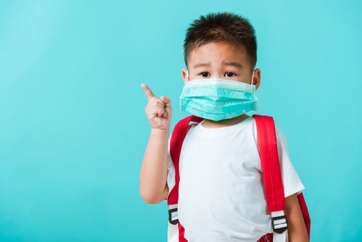 Back to school coronavirus Covid-19 education. Portrait Asian cute little child boy kindergarten wear face mask protective and school bag pointing finger to side, studio shot isolated blue background