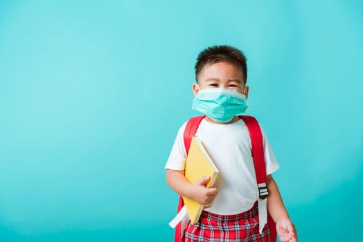 Back to school coronavirus Covid-19 education. Portrait Asian little child boy kindergarten wear face mask protective and school bag hold book before going to school, studio shot isolated background