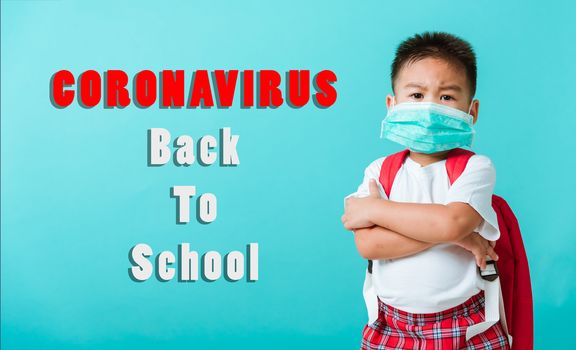 Back to school coronavirus Covid-19 education. Portrait Asian little child boy kindergarten wear face mask protective and school bag crossed arm before go school, studio shot isolated blue background