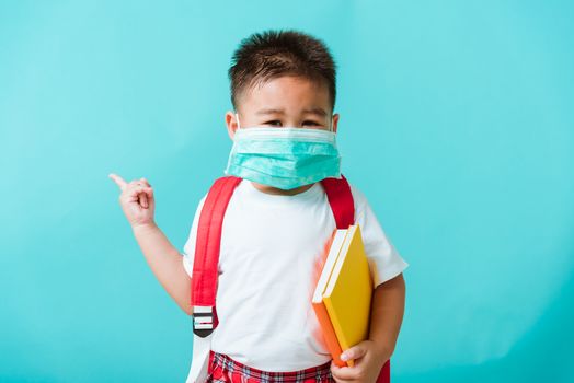 Back to school coronavirus Covid-19 education. Portrait Asian little child boy kindergarten wear face mask protective and school bag hold book before going to school pointing to side away, studio shot