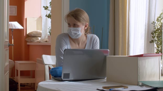Portrait of a young woman working from home. Lady in a protective mask and gloves at the kitchen table near computer. Remote job, quarantine. COVID-19 pandemic
