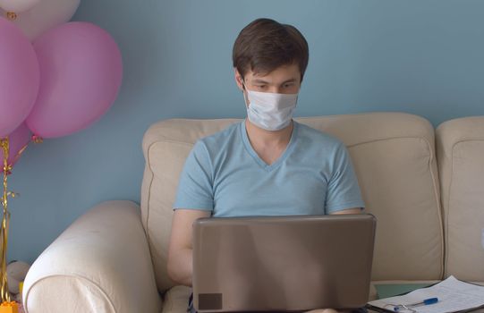 Home office. Man working remotely. Man in protective mask sitting on the sofa in bright room and typing in the computer. Quarantine, coronavirus epidemic.