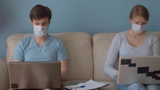 Home office. Couple on remote job. Man and woman in protective masks sitting on the sofa in bright room and typing in their computers. Quarantine, coronavirus epidemic.