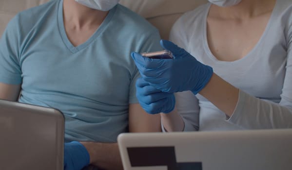 Close up female hands in protective blue gloves holding smart phone. The wife shows something to her husband. Quarantine, coronavirus epidemic.