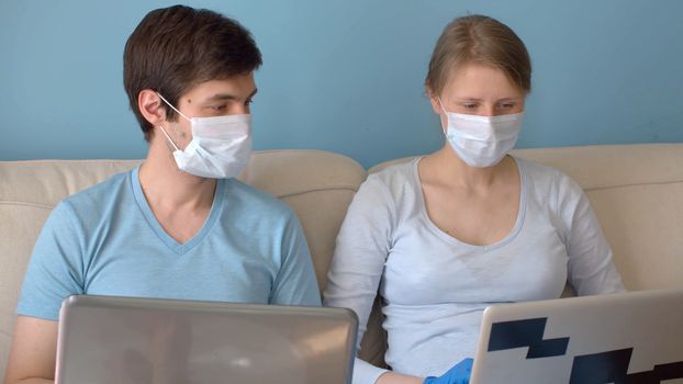 Home office. Couple on remote job. Man and woman in protective masks sitting on the sofa in bright room and typing in their computers. Quarantine, coronavirus epidemic.