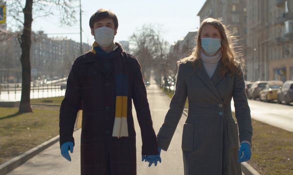 Beautiful couple in masks and blue latex gloves walking during coronavirus epidemic the city. Young blond woman and man holding hands, talking, smiling and rejoicing early spring sunny day. Covid-19