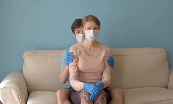 Man relaxing his wife by gently stroking her arms and shoulders. Nice couple in protective masks and gloves sitting on the sofa in bright room during coronavirus quarantine. Covid-19 stress