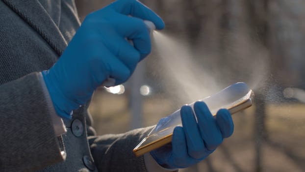 Close up woman's hands in protective gloves sanitaizing the smart phone outdoors. She sprays a sanitizer and the drops sparkle in the sun. Hygiene during coronavirus epidemic. Prevention of infection