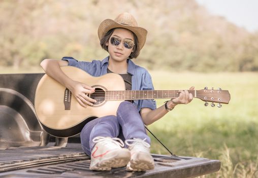 Young asian women short hair wear hat and sunglasses playing guitar ,sit on pickup truck in countryside Thailand