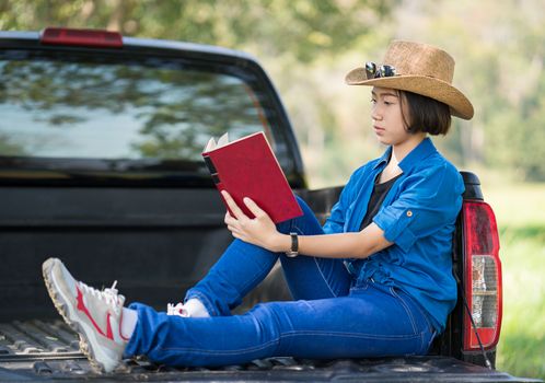 Young asian women short hair wear hat and sunglasses read a book ,sit on pickup truck in countryside Thailand