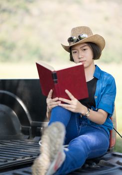 Young asian women short hair wear hat and sunglasses read a book ,sit on pickup truck in countryside Thailand