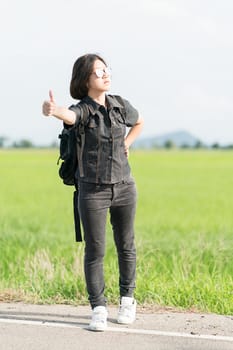 Young asian woman short hair and wearing sunglasses with backpack hitchhiking along a road in countryside Thailand