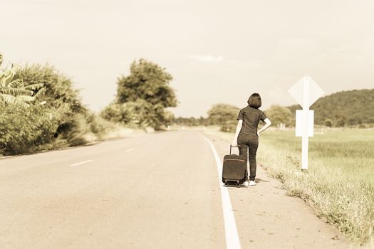 Young asian woman short hair and wearing sunglasses with luggage hitchhiking along a road in countryside Thailand