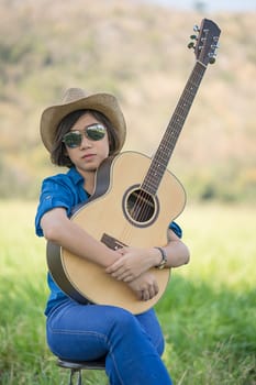 Young asian women short hair wear hat and sunglasses sit playing guitar in grass field countryside Thailand