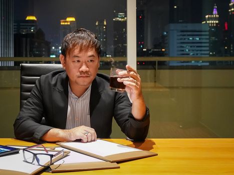 Asian businessman looking at liquor glass in his hand because he can't solve the financial problem. Meeting room on high floor at the night time. Bangkok's bustling downtown area.