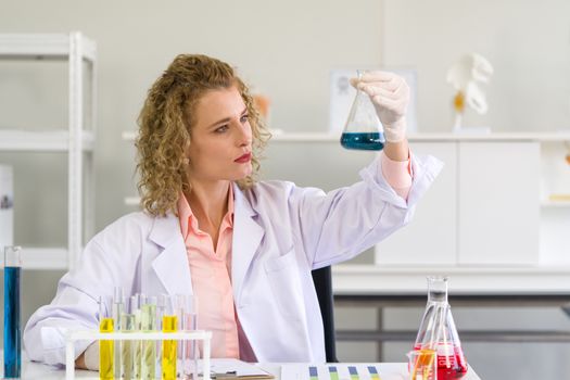 The young blonde scientist is considering the color of chemicals to observe the acidity of the alkalinity. Working atmosphere in chemical laboratory. Test tubes filled with chemicals on the table.