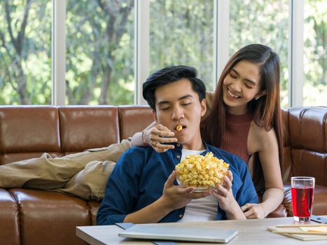 Young couples spend holidays in the living room. The young man was wearing comfortable clothes, showing his face, satisfied with the taste of afternoon snacks. Eating popcorn on the sofa.