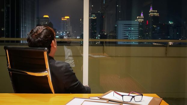 Asian businessman looking outside the window while review brief document after work. Meeting room on high floor at the night time. Bangkok's bustling downtown area.