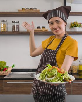 The housewife dressed in an apron and a hair cap, smiled and offered a salad bowl on the front. Morning atmosphere in a modern kitchen.
