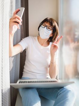 Pretty woman in medical mask works remotely from home. She is making selfie with Victory gesture on window sill with laptop on knees. Lockdown quarantine because of coronavirus COVID19. Self isolation at home.