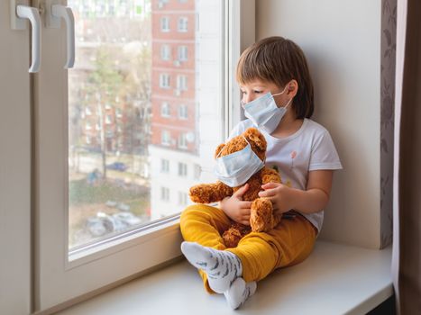 Toddler boy sits with teddy bear in medical masks. Kid with plush toy look through window outside. Browh plush bear and child on home quarantine because of coronavirus COVID-19.