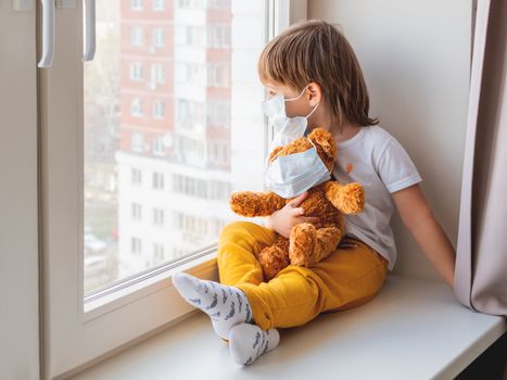 Toddler boy sits with teddy bear in medical masks. Kid with plush toy look through window outside. Browh plush bear and child on home quarantine because of coronavirus COVID-19.