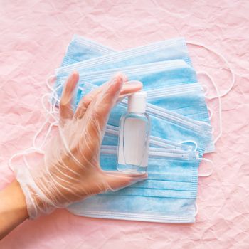 Top view on hand in protective glove with medical masks and sanitizer in transparent bottle. Woman doctor. Coronavirus COVID-19 concept on pink crumpled background.