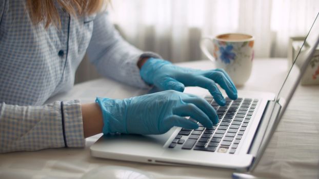 Close up female hands in protective blue gloves typing on a computer. Young woman working from home. Remote job, quarantine. COVID-19 pandemic