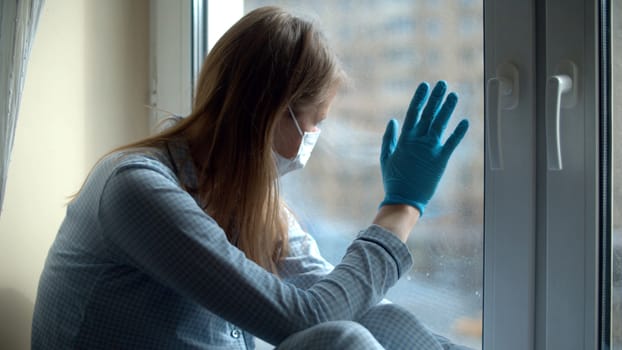 Young beautiful woman in home clothes and a protective mask sitting on the windowsill and looking out the window. Quarantine during the coronavirus epidemic. Covid-19 pandemic