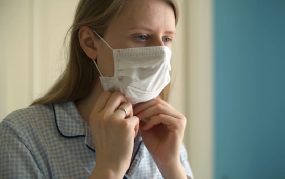 Close up portrait of a young woman putting on a protective mask in bright kitchen. Healthy and safety life concept. Covid-19 pandemic