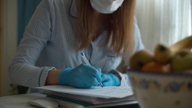 Young woman working from home. Lady in a protective mask and gloves writing something on the paper. Bright kitchen, bowl with fruits. Remote job, quarantine. COVID-19 pandemic