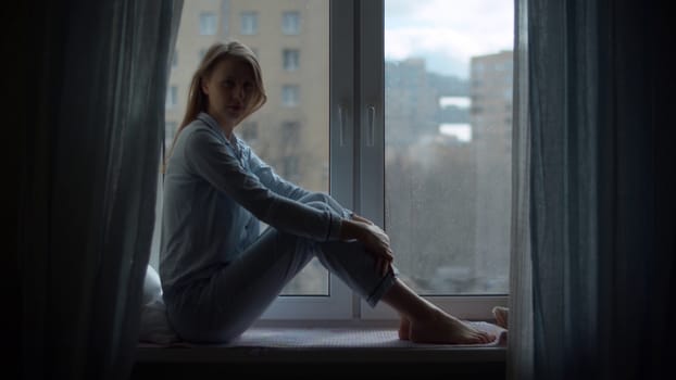 Young beautiful woman in home clothes and a protective mask sitting on the windowsill and looking out the window. Quarantine during the coronavirus epidemic. Covid-19 pandemic