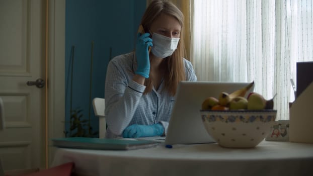 Portrait of a young woman working from home. Lady in a protective mask and gloves looking in a computer and talking by smart phone. Remote job, quarantine. COVID-19 pandemic