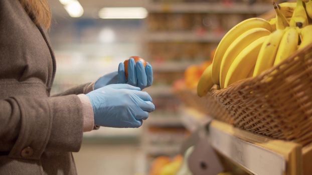 Close up hand of a woman in blue protective gloves choosing oranges in the supermarket. Coronavirus epidemic