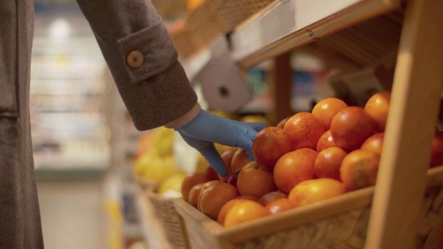 Close up hand of a woman in blue protective gloves choosing oranges in the supermarket. Coronavirus epidemic