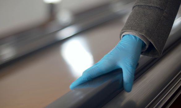 Close up female hand in protective blue glove holding subway escalator railing. Coronavirus epidemic in the city. Healthy and safety lifestyle concept. Covid-19 pandemic