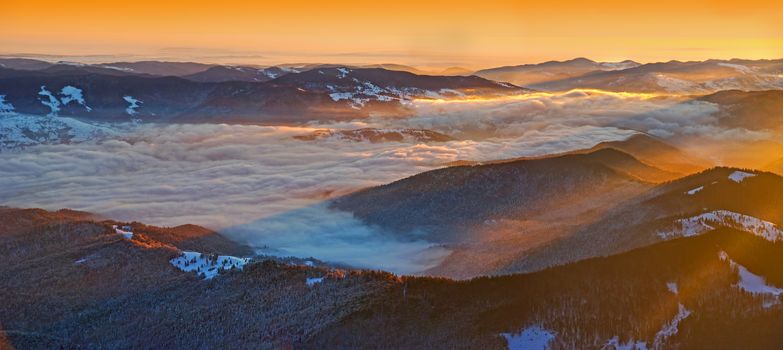 Winter sunrise panorama from the summit in Romanian Carpathians; misty valley and sun rays.